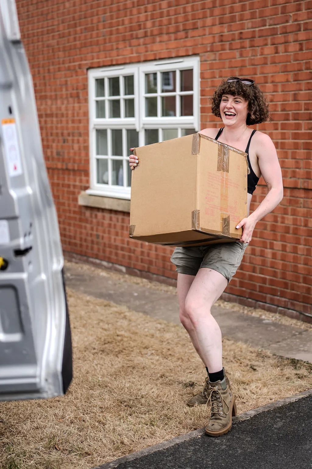 Hannah, founder of Han With A Van, laughing as she carries a large cardboard box past a brick house on a sunny moving day, with the back of a van beside her.